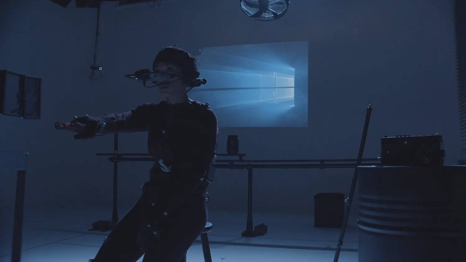 Performeuse assise en combinaison à marqueurs avec caméra faciale sur casque, dans un studio bleu